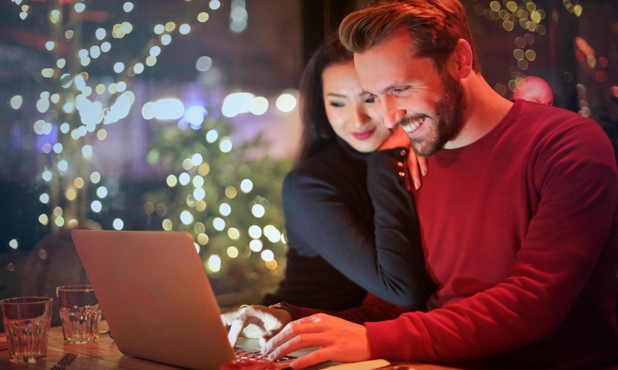 A couple using a laptop at a cozy restaurant with festive bokeh lights, enjoying online shopping together.